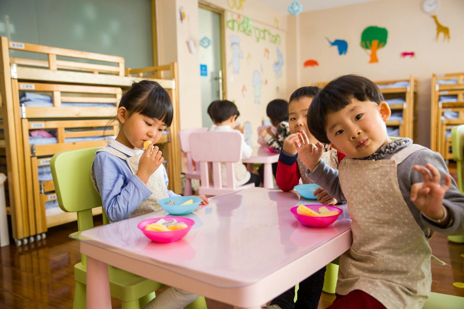 Home Kids seated around a table in a colorful classroom, eating snacks happily.