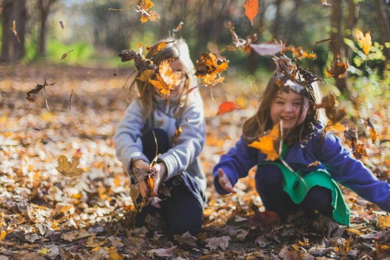 Home Two smiling girls playing joyfully amidst colorful fall leaves in a sunlit forest